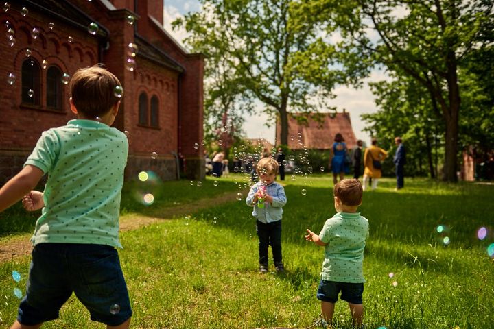 Kinder spielen auf diesem Hochzeitsfoto mit Seifenblasen auf einer Wiese an einer Kirche.