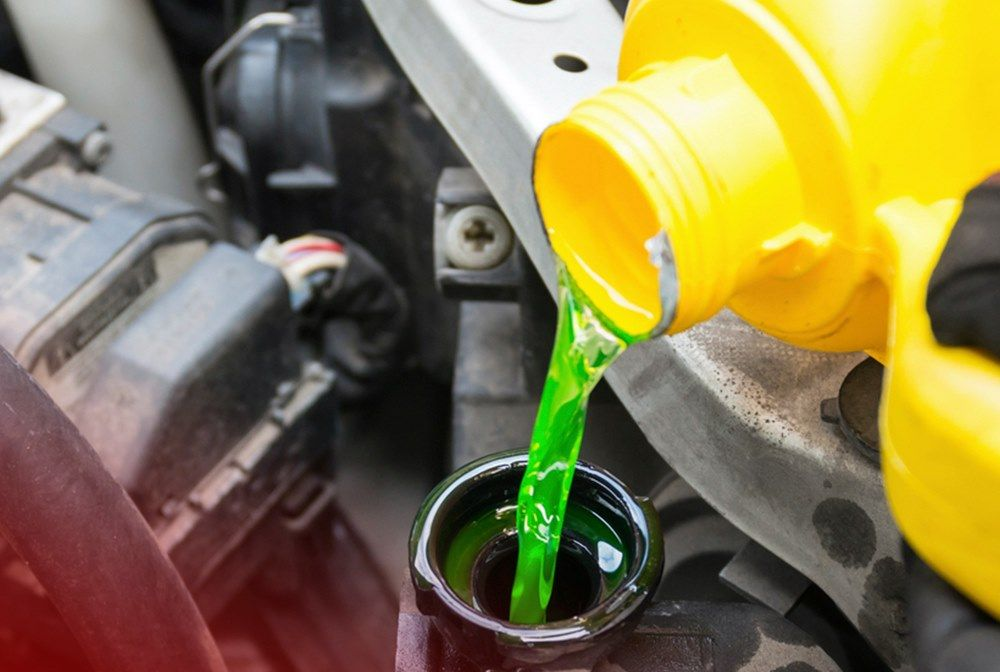 A person pours green coolant into a car's radiator from a yellow jug, engine bay.