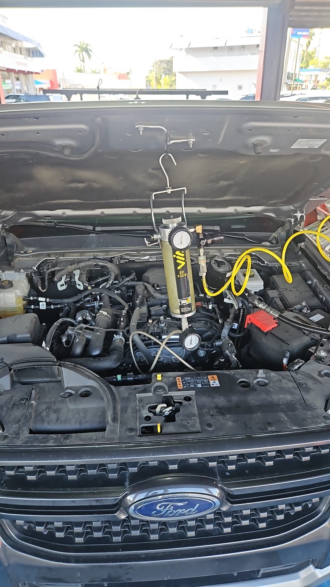 Car engine bay open with a fluid-extraction tool in use. Ford logo on the grill.