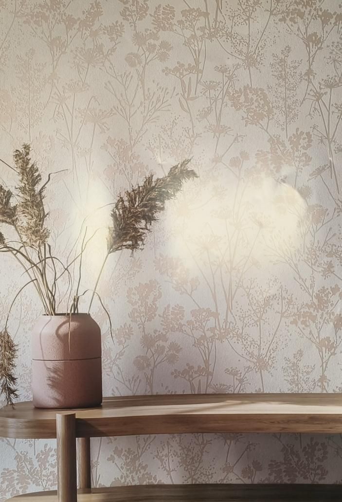 Un vaso con piante secche su uno scaffale di legno davanti a una carta da parati floreale beige.