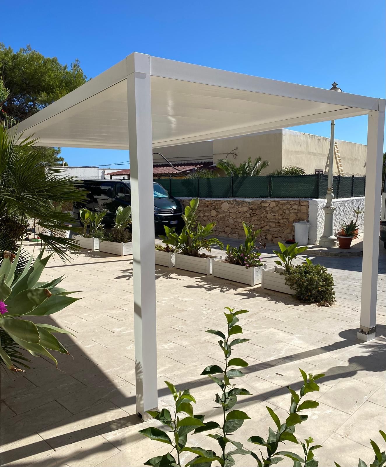 Pergola bianca su un'area pavimentata, con piante e una casa sullo sfondo sotto un cielo azzurro.