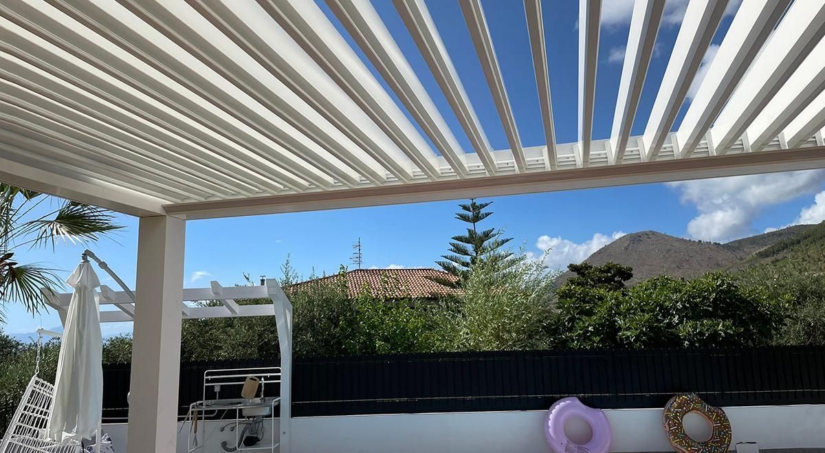 Tetto bianco a pergola con doghe aperte, che si affaccia su un paesaggio verde con cielo azzurro e montagne.