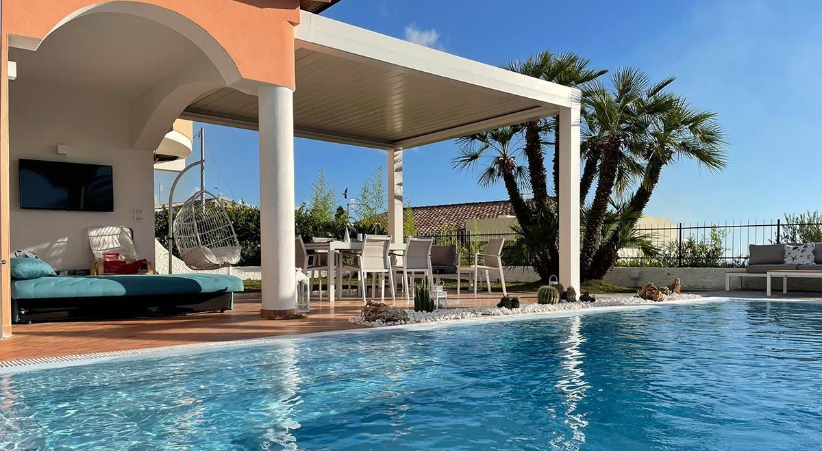 Patio a bordo piscina con tavolo e sedie sotto un pergolato bianco, con vista sulla piscina blu.