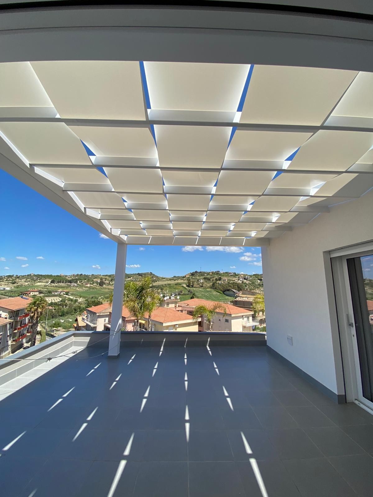 Pergola con ombrelloni quadrati bianchi che proiettano ombre su una terrazza panoramica piastrellata; vista sulla città e sul cielo azzurro.