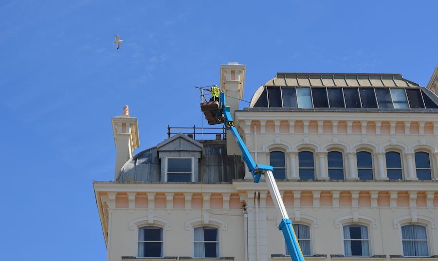 Person in a lift working on a white building's roof. Blue lift and sky. Seagull flying above.