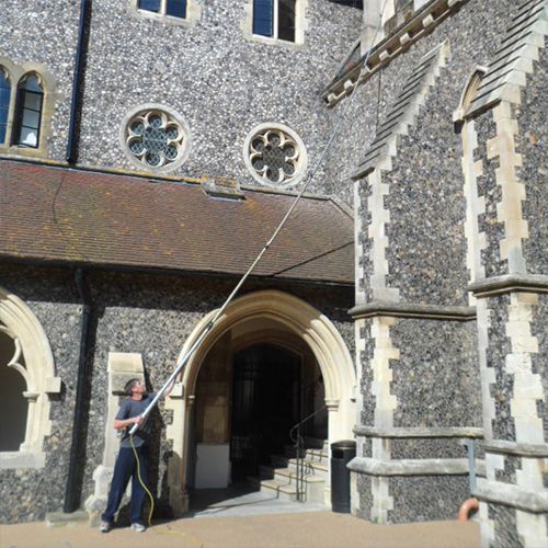 Man using a long pole to clean windows on a stone church. Blue sky, sunlight.