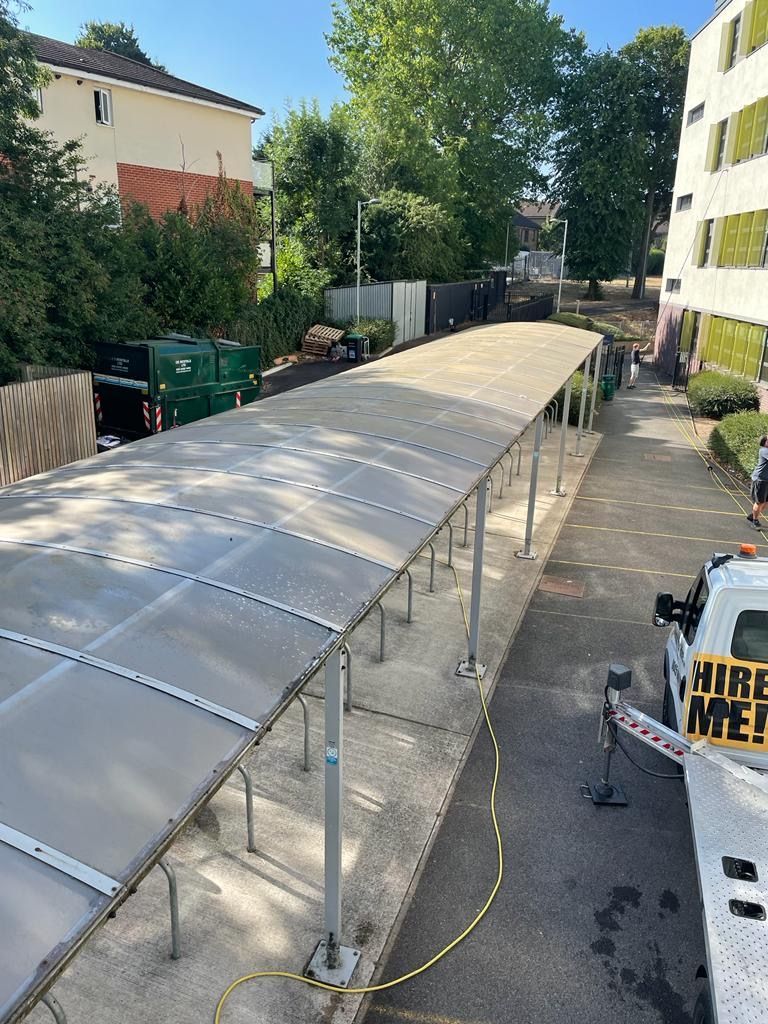 Bike shelter with a clear roof, next to an apartment building. Green bins and a truck are visible behind it.