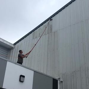 Man on rooftop cleans the side of a metal building with an extended brush under a cloudy sky.