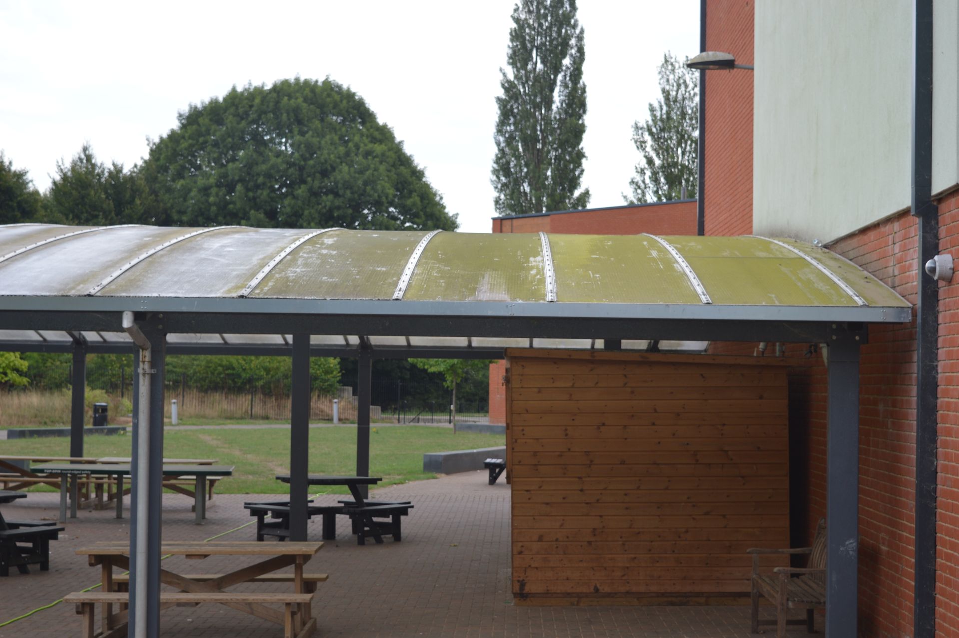 Picnic tables under a covered patio next to a brick building. Green trees and sky visible.