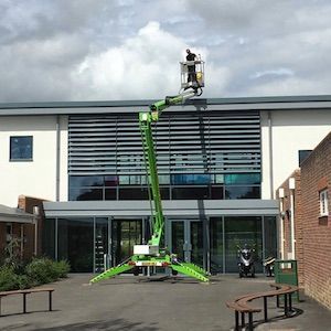 A green lift crane with a worker in the basket is parked near a building with glass windows.