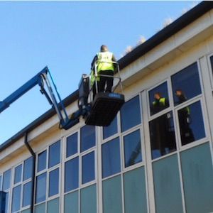 A worker in a lift basket cleaning gutters on a building with multiple windows.