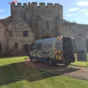 Van parked in front of castle, doors open, hose running. Green grass, gray stone building with flag.