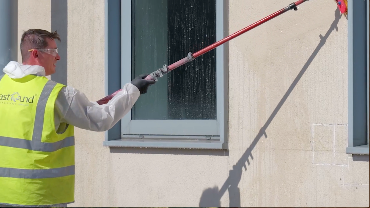 Person in safety gear cleaning a building's exterior with a long-handled brush.