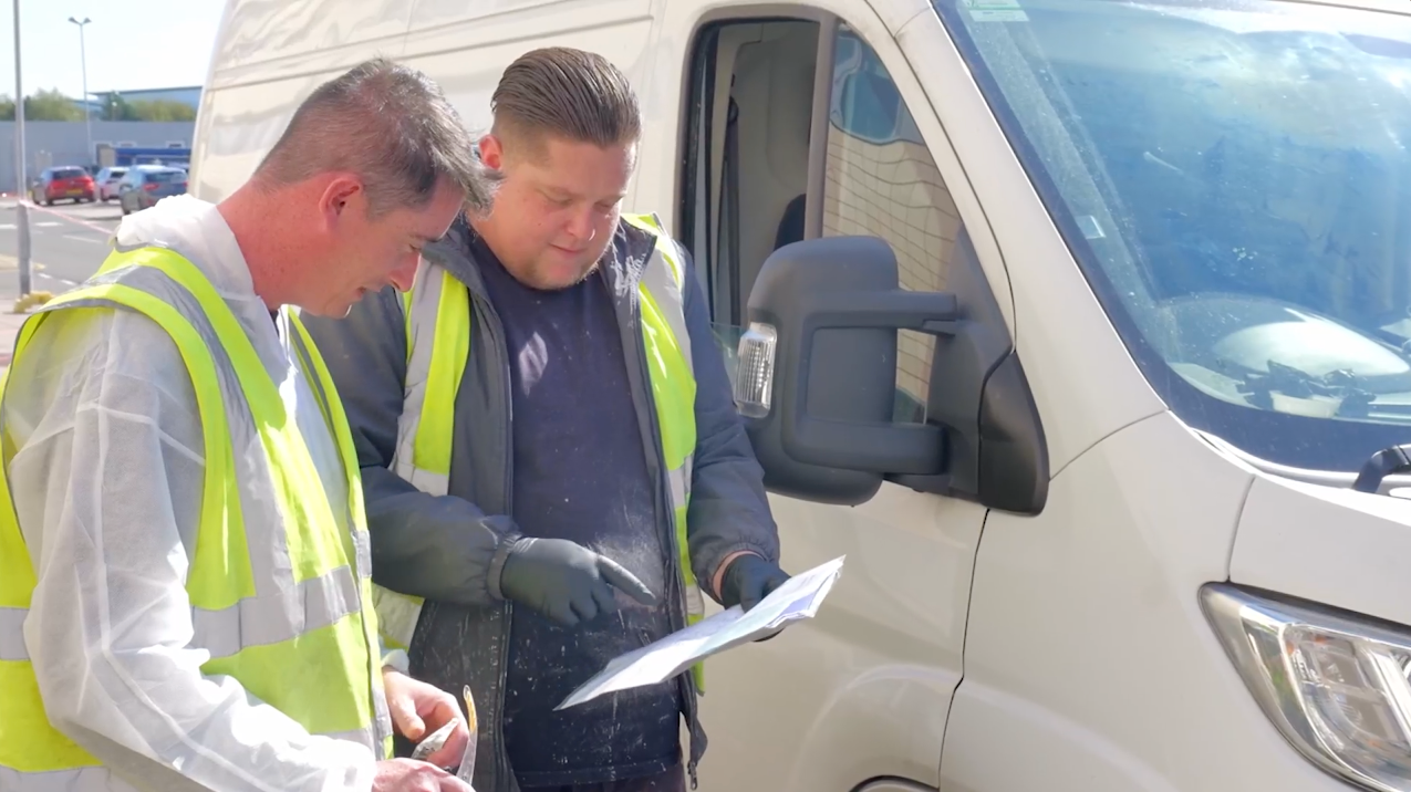 Two men in high-vis vests reviewing paperwork next to a white van outdoors.