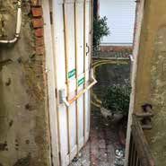 A white door propped open with a metal bar.  Brick and weathered walls, a small outdoor area visible beyond the door.
