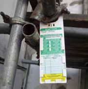 Scaffolding with inspection tag, green and white, attached near metal pipe.