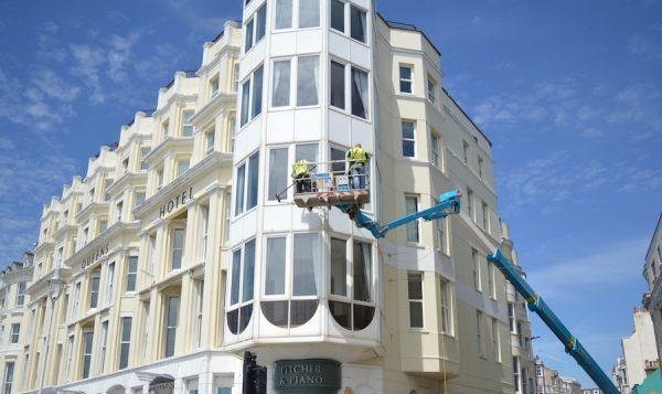 Workers in a lift repairing a tall white building with many windows, on a sunny day.