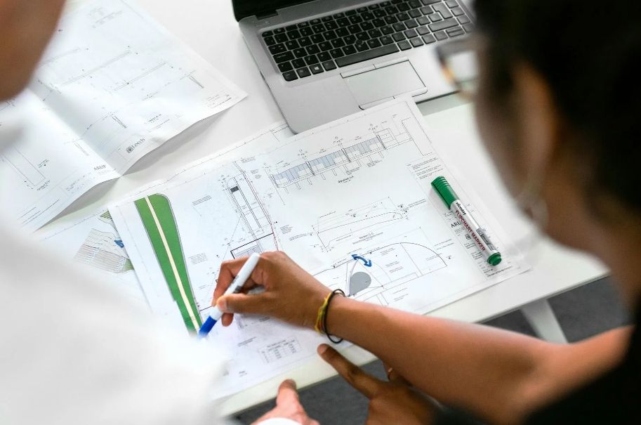 Two people reviewing architectural plans with laptop and markers on a desk.