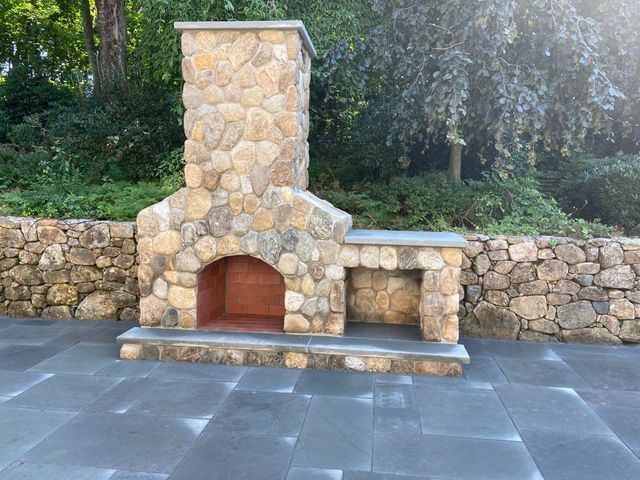 A stone fireplace is sitting on a patio next to a stone retaining wall.