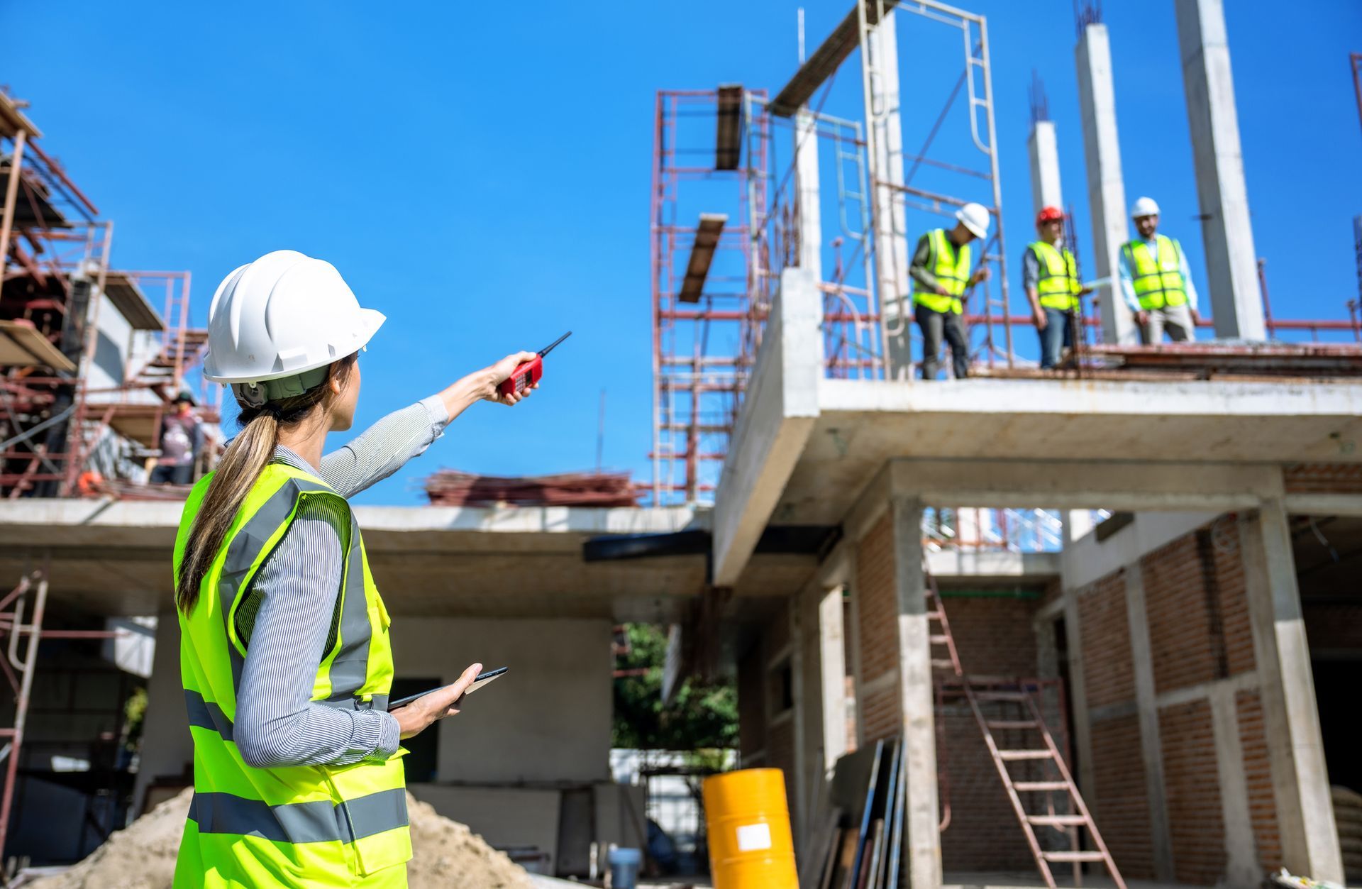 Construction worker in white hard hat and neon vest directs workers on a building site.