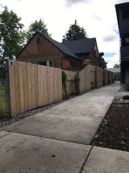 Wooden fence alongside a concrete path, leading to a house with a dark roof.