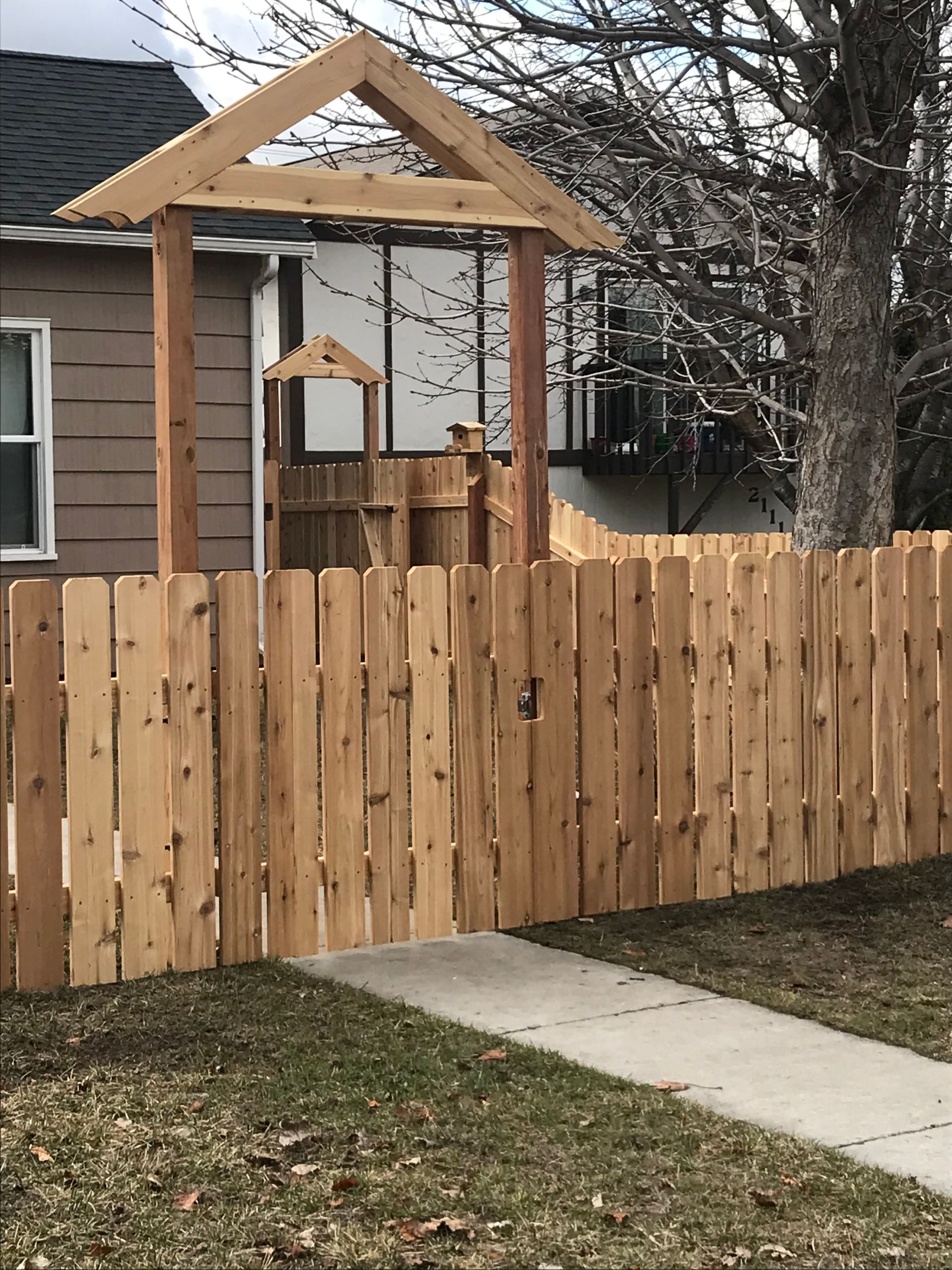 Wooden fence with gate in front of a playground structure, in front of a house and sidewalk.