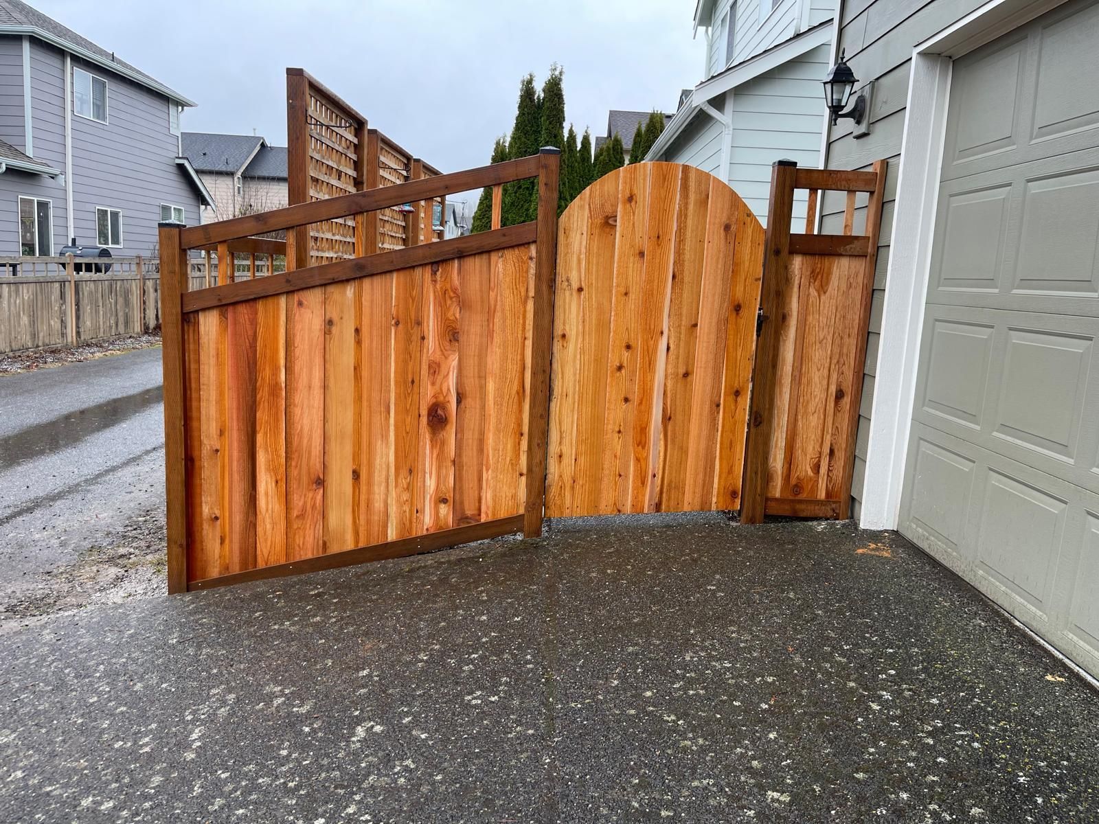 Wooden gate and fence with an arched gate section, set on a wet driveway, next to a garage.