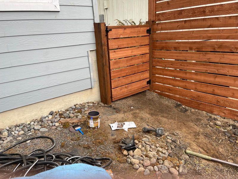 Wooden gate and fence section attached to a light blue house. Construction materials scattered on the ground.