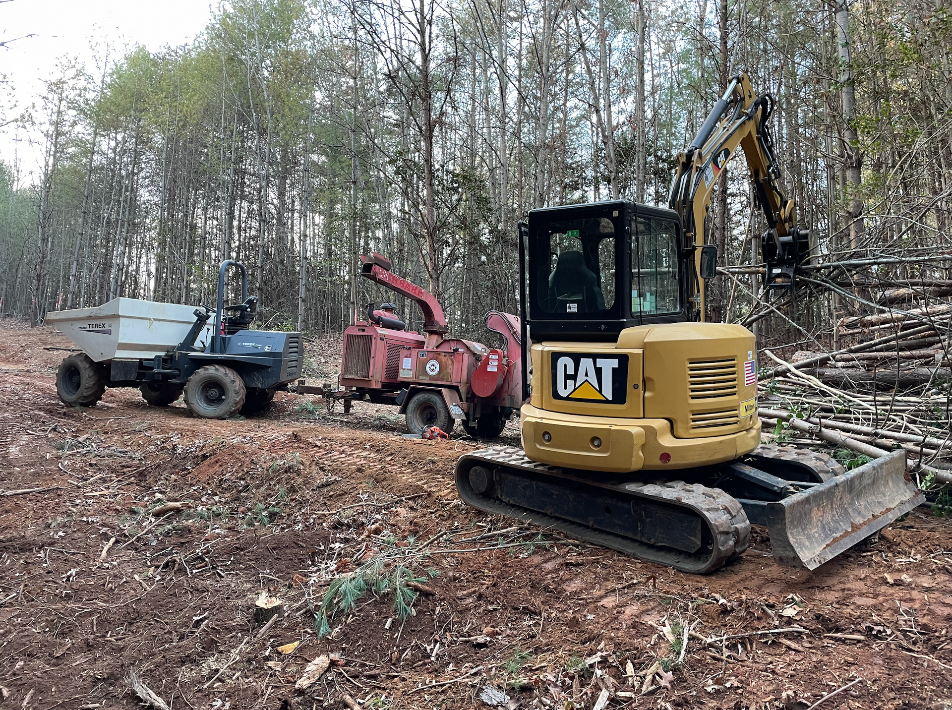 A cat excavator is sitting in the middle of a forest next to a truck.