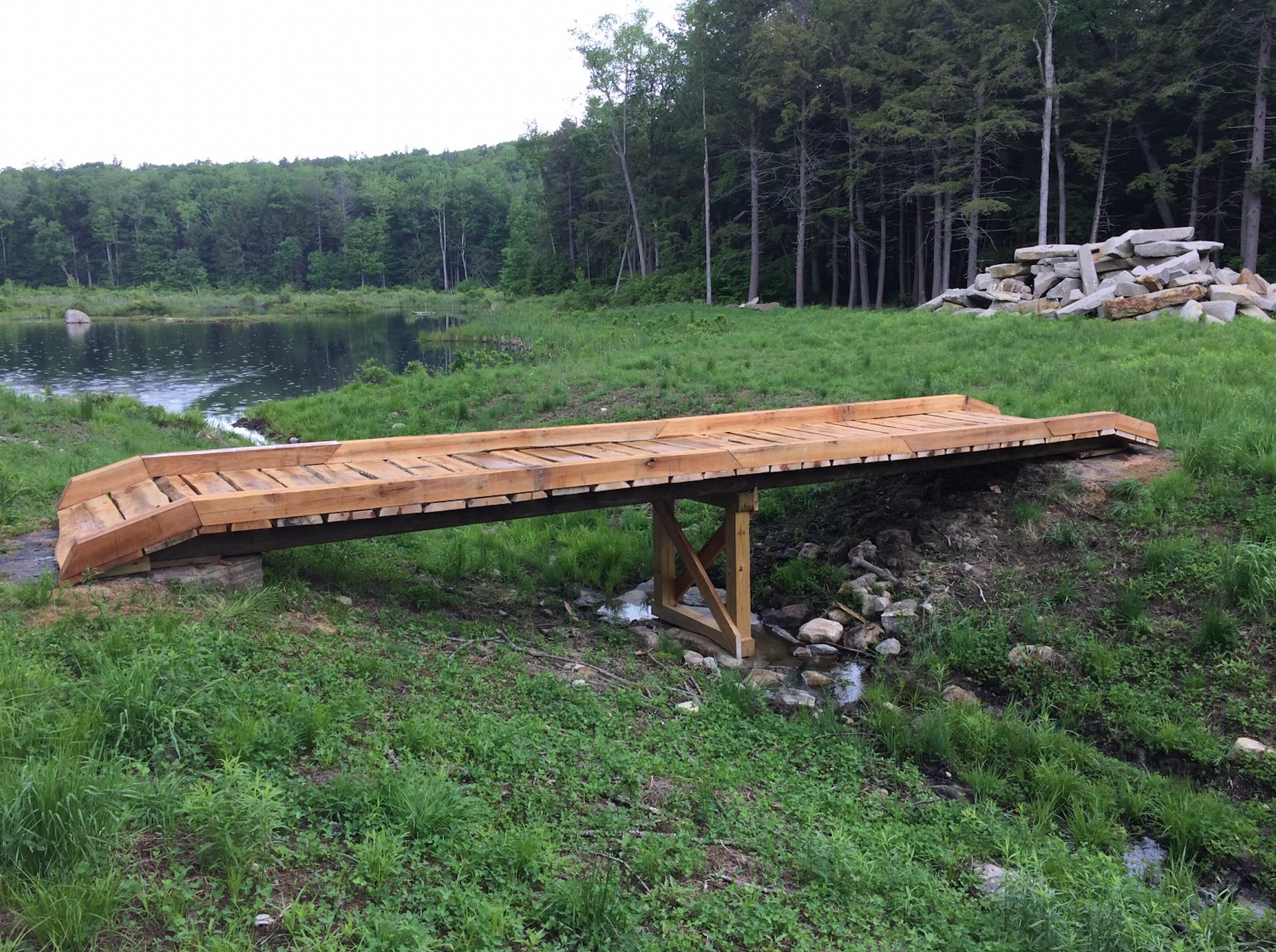 A wooden bridge is sitting in the middle of a grassy field next to a body of water.