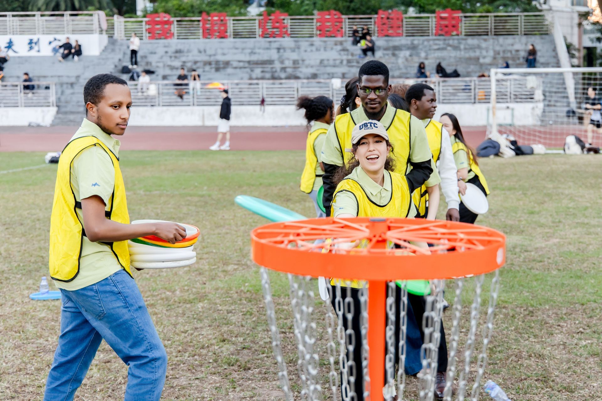 People playing disc golf in a park, wearing yellow vests, with an orange basket, and a stadium in the background.