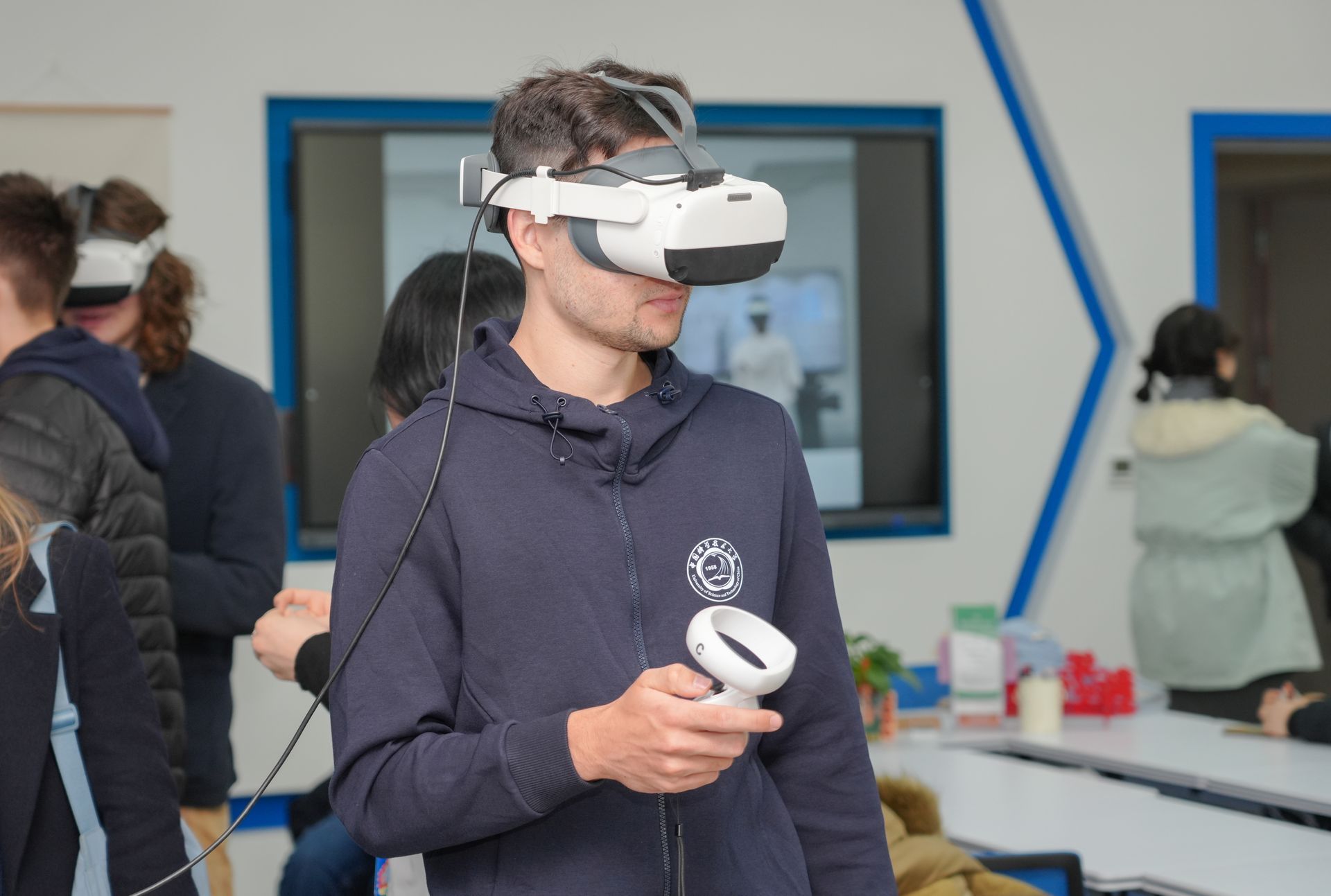 Man wearing VR headset and holding a controller indoors, others with headsets in the background.