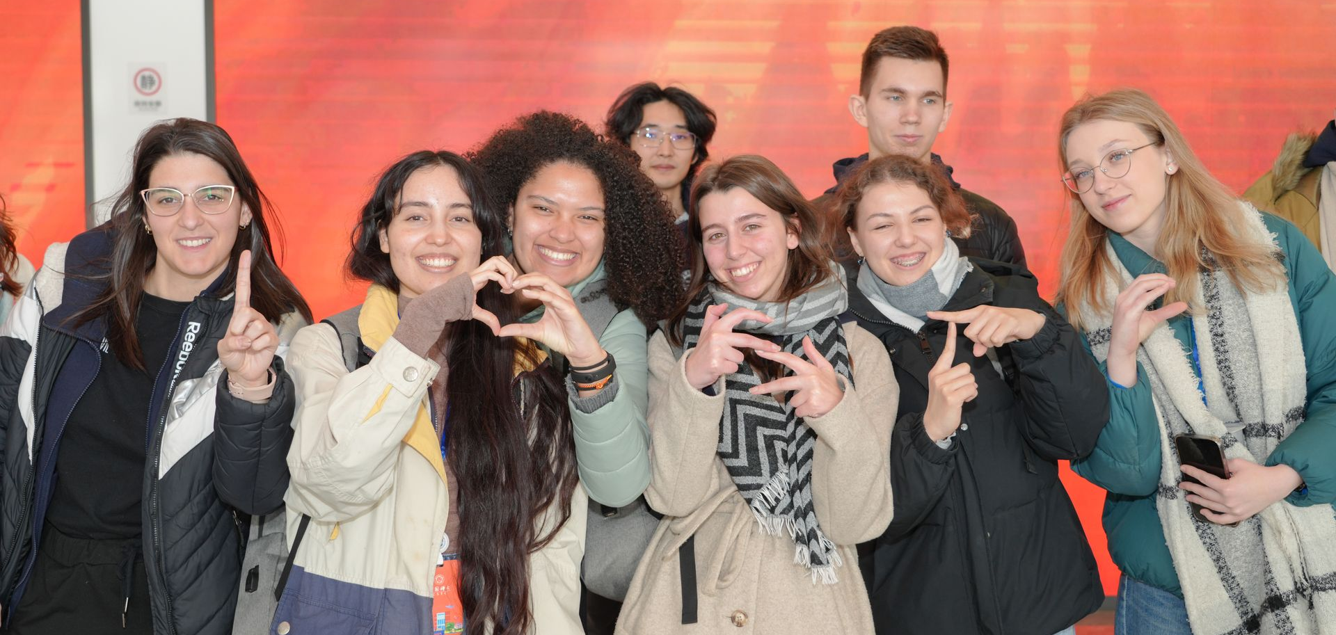 Group of people making heart and 'hang loose' hand gestures, smiling, posing in front of an orange background.