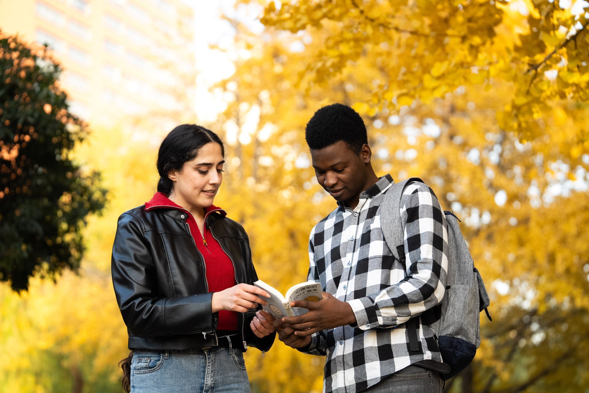 Two people looking at a book outdoors, surrounded by yellow leaves.