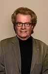 Man with glasses, tan jacket, black turtleneck, smiles slightly.