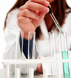 Scientist in lab coat uses dropper to add liquid to a test tube with green liquid.