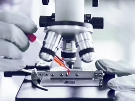 Hands in gloves add red liquid to a microscope slide under a microscope. Lab setting.