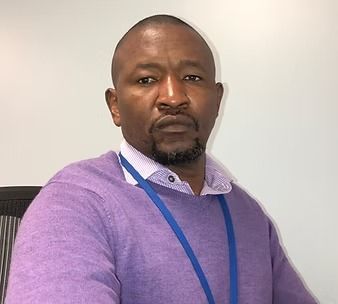 Man with a short haircut wearing a purple sweater, blue lanyard, seated in an office.