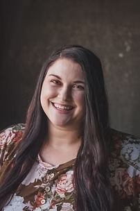 Woman with long dark hair smiling, wearing a floral top, against a muted brown background.