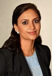 Woman in a blazer smiling, with dark hair and a light blue shirt, in a studio setting.