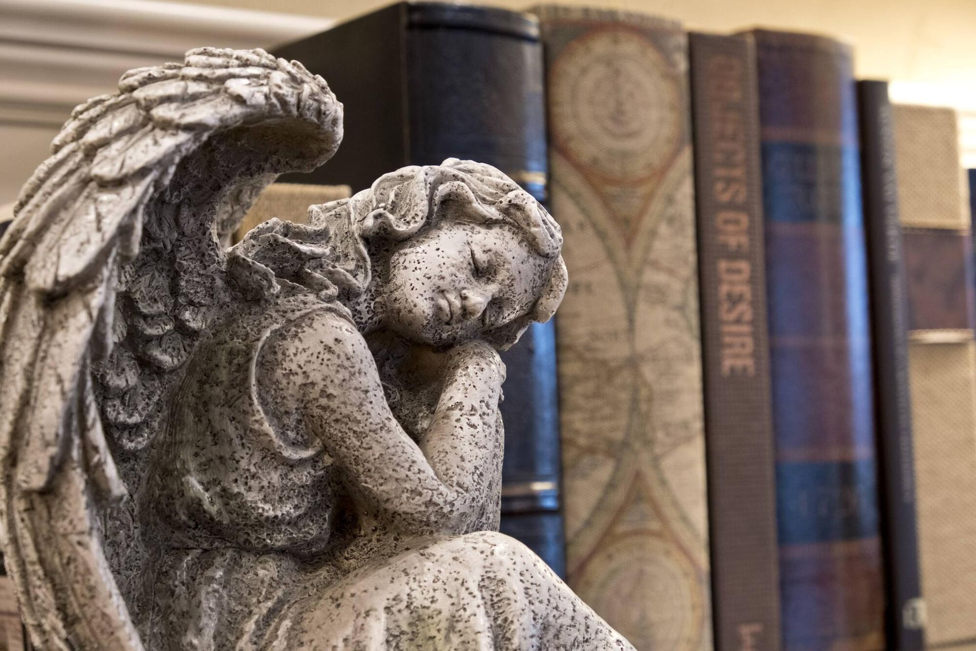 Stone angel figurine next to a row of books; cream-colored, resting head on hands.