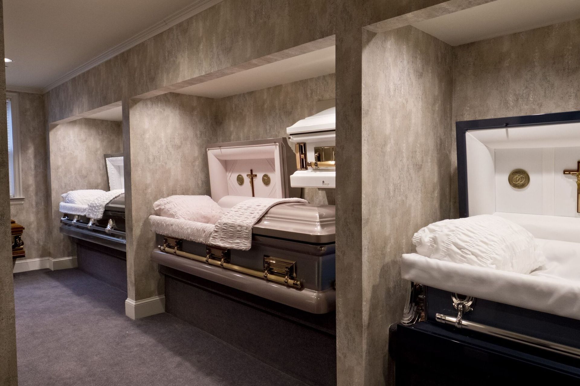 Display of several open caskets in a funeral home showroom, each in a separate alcove, with various colors and designs.