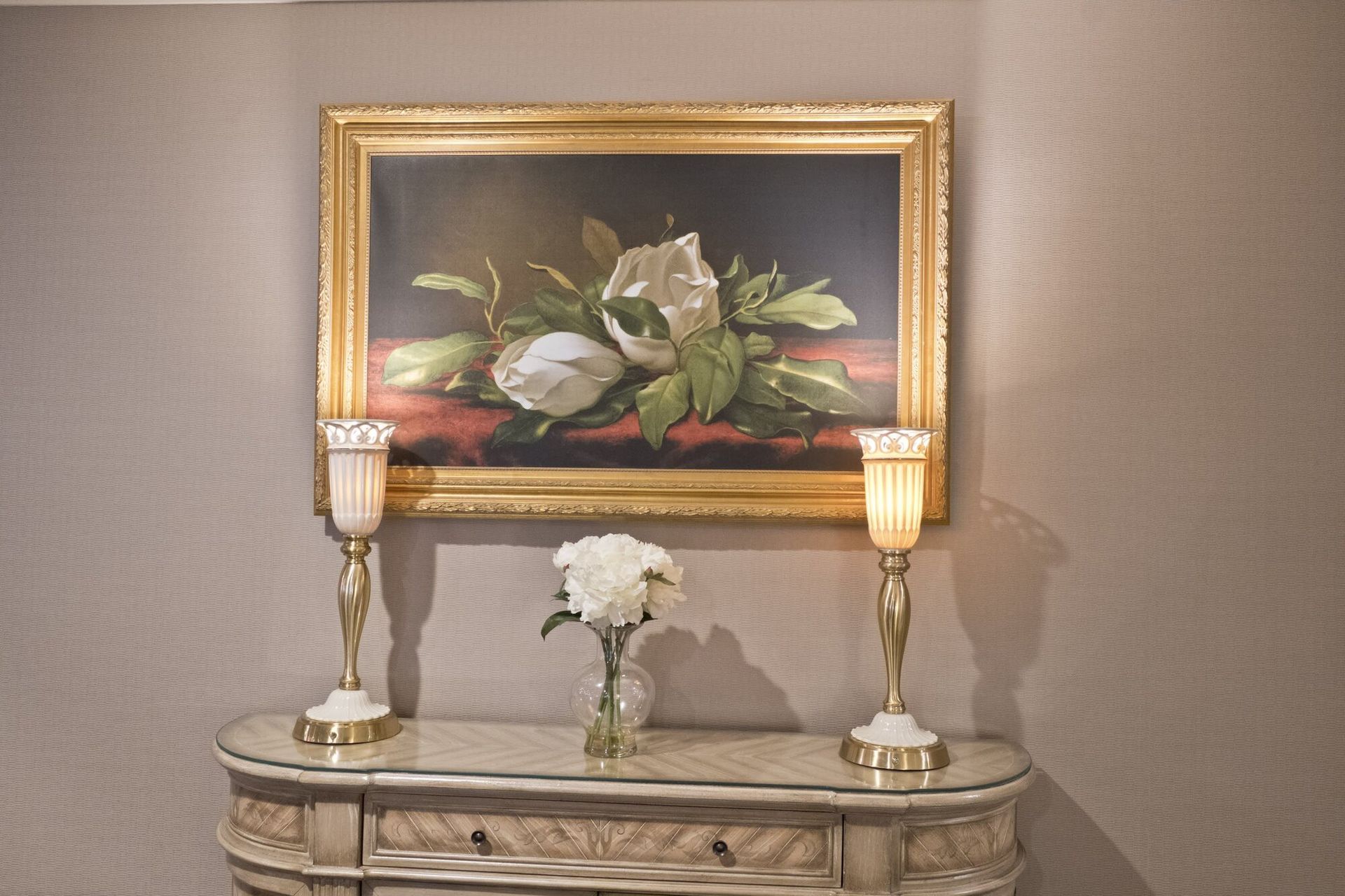 Oil painting of white flowers and greenery above a console with lamps and vase.