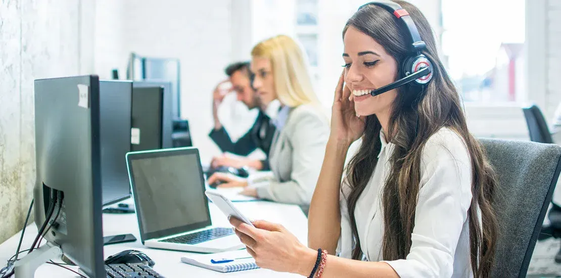 Mujer con auriculares sonriendo, sosteniendo un teléfono, trabajando en una oficina con otras personas en computadoras.