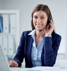 Mujer con auriculares, sonriendo, trabajando en un escritorio con portátil y papeleo. Traje azul, en un entorno de oficina.