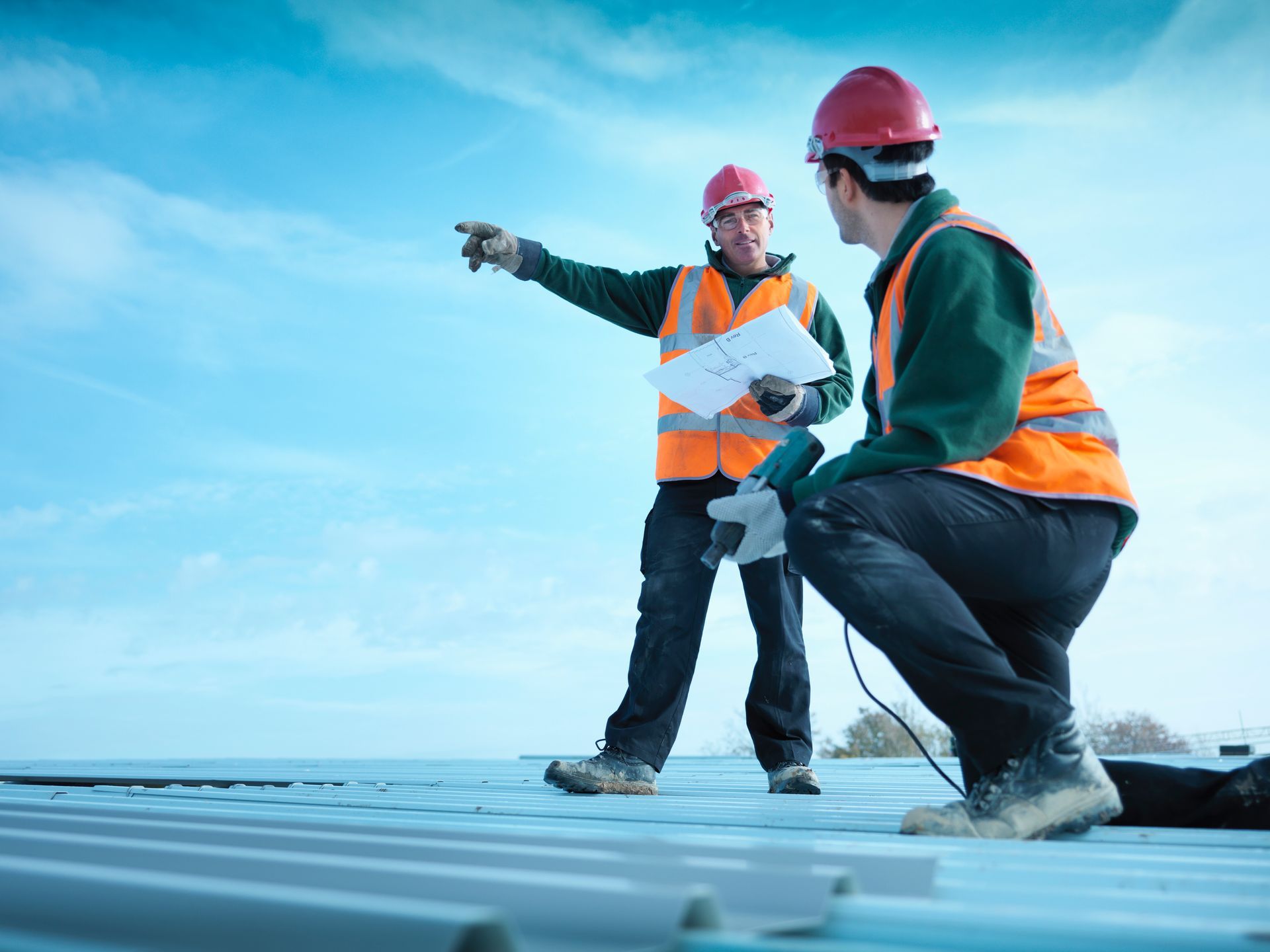 Two construction workers are working on a roof and one is pointing at something.
