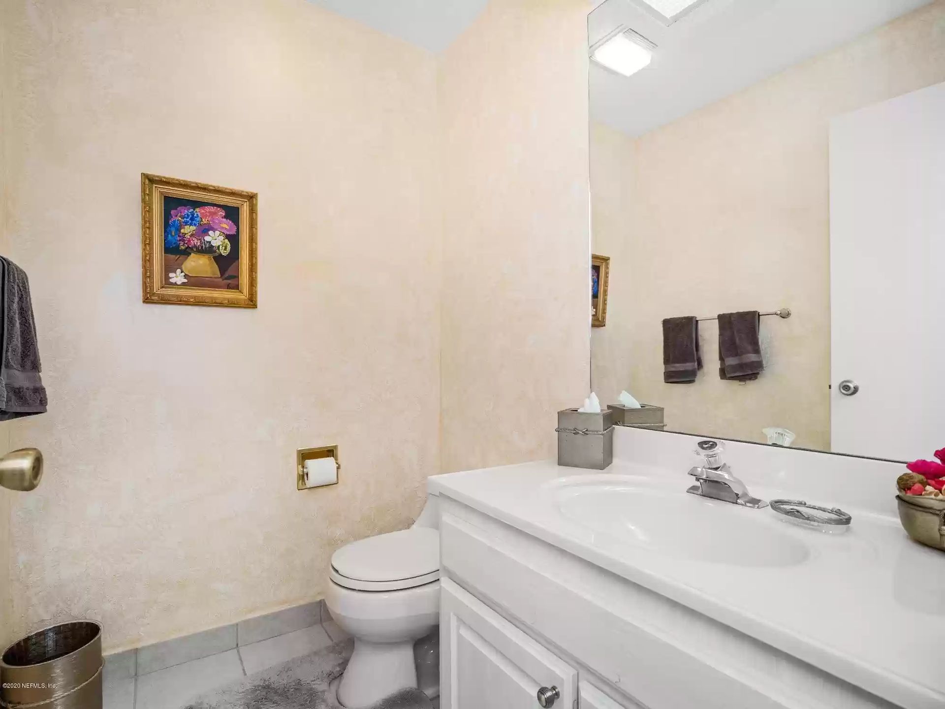 Powder room with white vanity, toilet, art, and mirror; beige walls, gray tile floor.