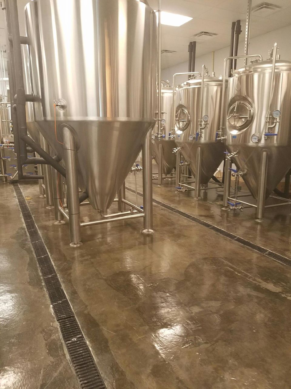Stainless steel brewing tanks in a brewery, with a shiny floor and drainage channels.