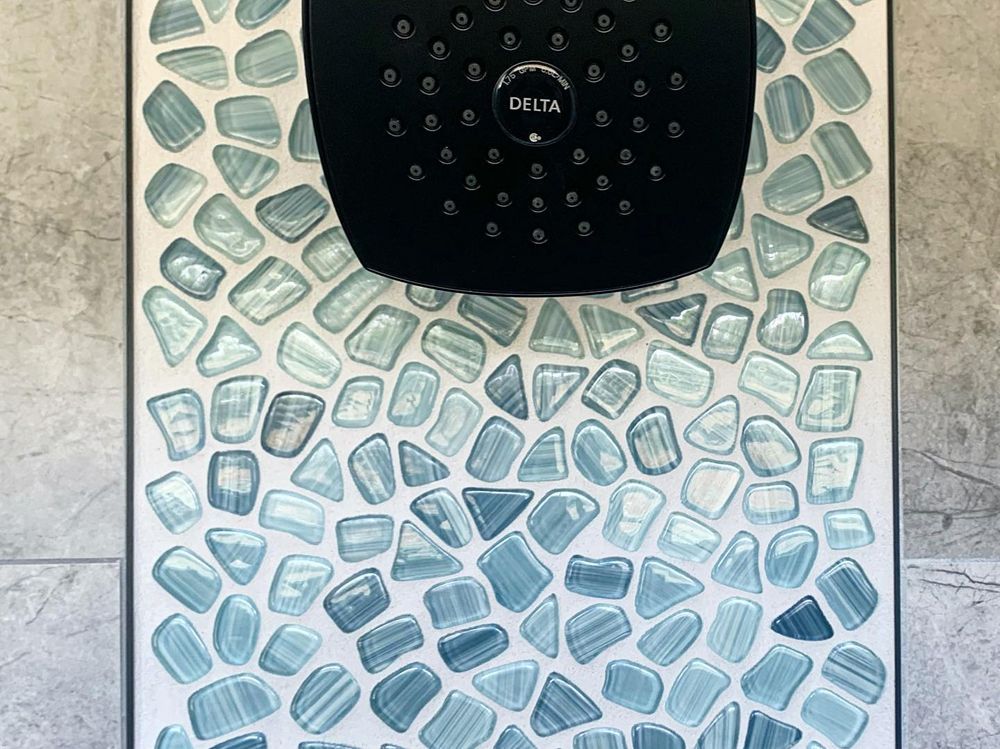 Black showerhead over mosaic tile in shades of blue and white on a gray wall.