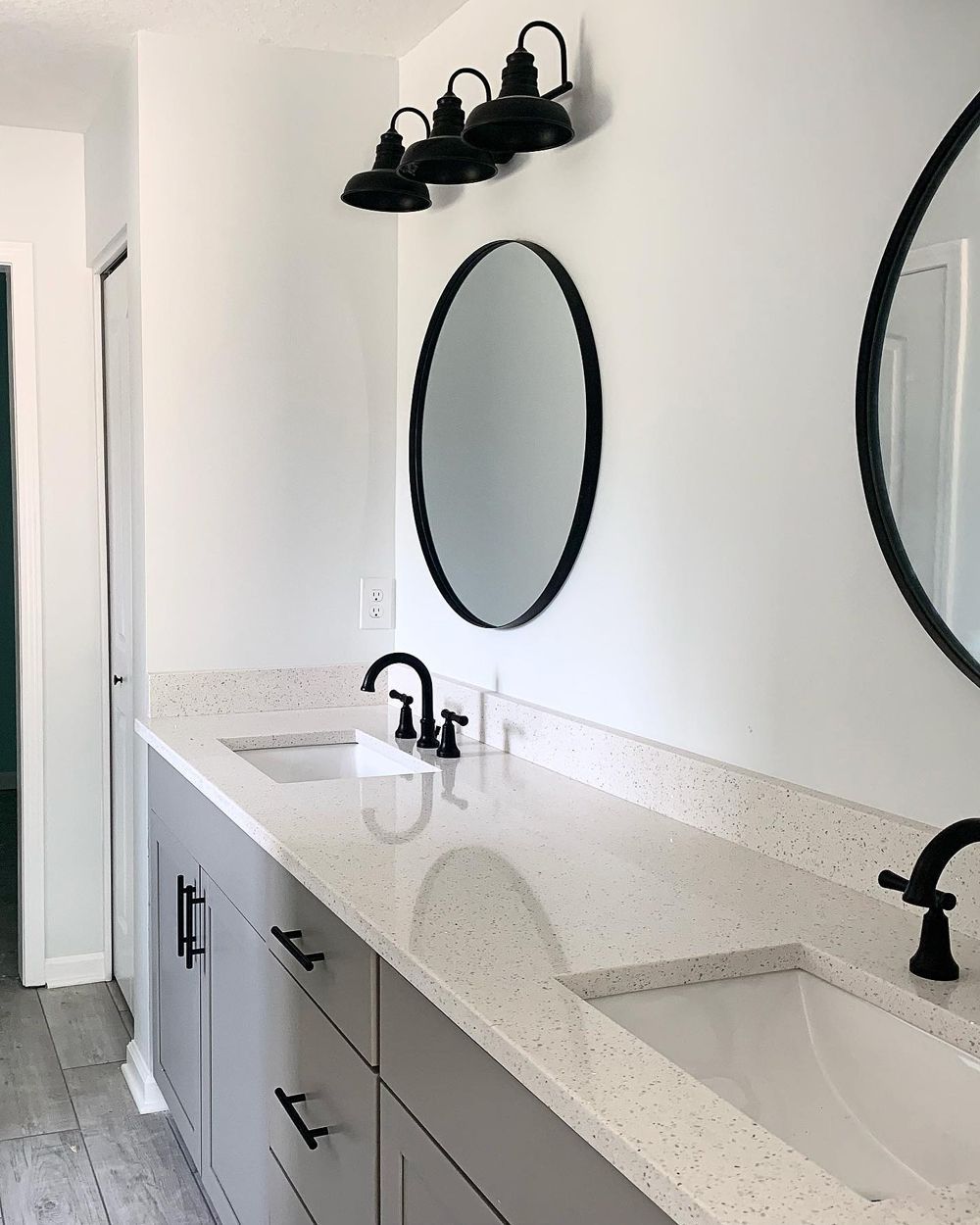 A modern bathroom with a gray vanity, white countertop, black fixtures, oval mirrors, and gray floor.